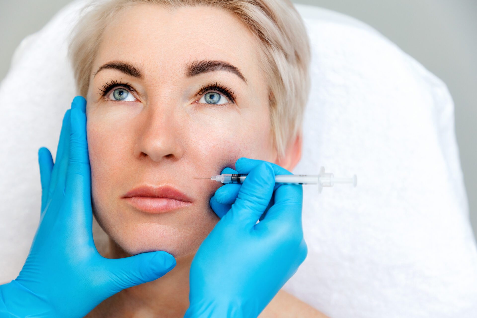 Beautician in medical gloves with syringe injects filler in nasolabial fold. Close-up portrait of beautiful adult woman getting injection in the cosmetology salon.